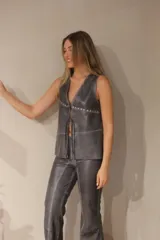 Chaleco marrón de cuero con cuello en V, cierre frontal con broches metálicos y detalle de tachas en el borde. Diseño con paneles y flecos en la parte inferior.