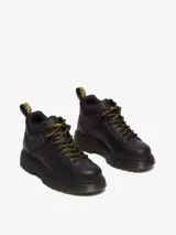 Botín de cuero negro Dr. Martens modelo Woodard, con cuello acolchado, cordones de tipo excursionista en amarillo y negro, suela acanalada Club Wedge con pespunte negro y tirador trasero con logo.