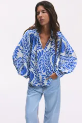 Blusa oversized con cuello elevado fruncido, abertura en V adelante y bajo recto. Mangas globo largas de corte raglán con abertura y botón en los puños. Estampado floral en tonos azules y blancos.
