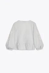 Blusa corta color celeste con textura arrugada, escote en V con puntilla, lazo ajustable en el frente y mangas 3/4 abullonadas con bordados florales a contraste.