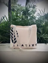 Bolso tipo tote bag de lona color crudo con asas largas de hombro. Estampado lateral con diseño de hoja y texto 'Nativo' en color negro.