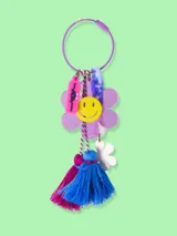 Llavero con aro metálico violeta y dijes colgantes: flor lila con cara sonriente amarilla, flor blanca, borla de hilo azul y borla de hilo fucsia.