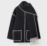 Conjunto de saco y bufanda color gris oscuro con detalles de costura en blanco. El saco es de corte holgado, con cuello alto, cierre frontal con botones, bolsillos frontales y mangas largas. La bufanda es larga y con flecos.