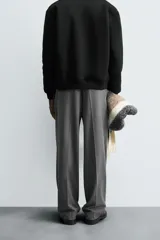 Pantalón gris de corte ancho con cintura elástica, pliegues frontales, bolsillos laterales y traseros de vivo. Incluye detalle de hebilla metálica en la trabilla y cierre frontal con cremallera y botón.