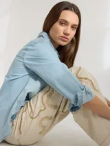 Camisa celeste de denim suave, corte relajado, con cuello clásico, cierre frontal con botones marrones, manga larga con puños abotonados, dos bolsillos de parche en el pecho y ruedo redondeado.