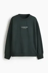 Buzo de tejido de punto color verde oscuro, con cuello redondo, hombros caídos y puños y bajo acanalados. Presenta un pequeño estampado centrado en el pecho con el texto "CANNES 1980" y debajo "La Côte D'Azur".