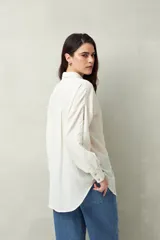 Camisa blanca de lino con botones de nácar, corte recto, cuello clásico y mangas largas con puños abotonados. Presenta detalles de calados tipo vainillas en el frente y las mangas.