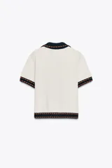 Chaqueta color blanco de punto con cuello solapa y manga corta. Tiene cierre frontal con botones color naranja y ribete en azul marino y naranja.