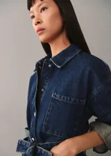 Chaqueta vaquera azul con cuello camisero, cierre frontal con botones metálicos, tres bolsillos delanteros, mangas largas con puños abotonados y cinturón de lazo.