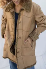 Abrigo gamulán marrón con cuello, solapa, puños y borde frontal de piel sintética blanca. Tiene cierre con botones y bolsillos laterales.