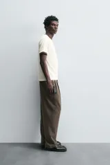 Pantalón negro de corte ancho con cintura elástica, pliegues frontales, bolsillos laterales y traseros de vivo. Tiene detalle de hebilla metálica en la trabilla y cierre frontal con cremallera y botón.