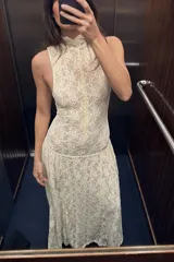 Vestido largo de encaje color blanco roto, con cuello alto, sin mangas y corte evasé. Incluye forro interior y cierre en la espalda.