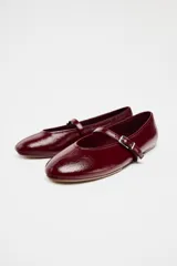 Zapato tipo bailarina color borgoña con efecto acharolado, acabado en punta redonda y tira con hebilla ajustable en la parte delantera.