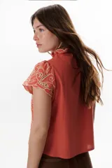 Blusa color coral o terracota con bordado calado floral en tono beige claro. Presenta cuello alto con volado y mangas cortas abullonadas.