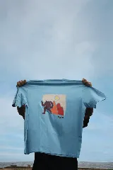 Remera celeste de manga corta con estampa de una figura humana marrón, montañas rojas y un sol amarillo.