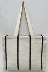 Bolso tipo tote tejido a mano, color crudo con rayas verticales negras y flecos en los laterales.