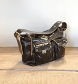 Bolso tipo hobo de cuero color marrón oscuro con acabado brillante. Tiene correa de hombro ajustable con hebilla dorada y múltiples bolsillos externos con cierre de broche metálico.