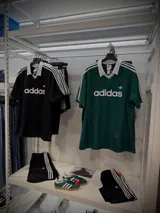 Jersey deportivo Adidas Originals de corte regular, color negro con textura de rayas verticales sutiles. Presenta cuello tipo polo en V con ribete blanco, tres rayas blancas en las mangas y el logo Trefoil bordado en blanco en el pecho.