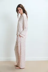 Pijama de dos piezas, compuesto por una camisa de manga larga y pantalón largo, ambos en tejido de viscosa color blanco con estampado integral de corazones rojos. La camisa tiene ribetes rojos en cuello, puños y abertura frontal. El pantalón tiene un fajín ancho color rojo liso.