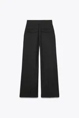 Pantalón negro de tiro alto con trabillas, doble pliegue, bolsillos laterales y tapetas en la espalda. Tiene pernera ancha y cierre frontal con cremallera y gancho metálico.
