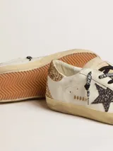 Zapatillas Golden Goose modelo Super-Star, de cuero color blanco con detalles en dorado glitter en el talón y estrella lateral negra con glitter. Cordones negros con texto estampado.