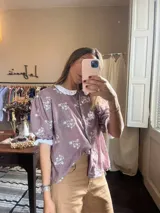 Camisa color lila con estampado de flores blancas, cuello bebé y mangas abullonadas con terminación de puntilla. Cierre frontal con botones.