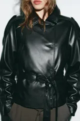 Chaqueta negra de efecto piel con cuello solapa, manga larga y trabillas en los hombros. Presenta bolsillos delanteros y cierre frontal con botones, ajustada a la cintura con un cinturón del mismo tejido.