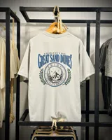 Remera de manga corta, corte oversized, color gris ceniza, con un estampado gráfico en la espalda que incluye texto "I Hiked The Great Sand Dunes", la palabra "Tribu", y un diseño circular con montañas, árboles y la frase "Quality Brand".