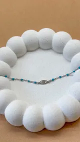 Pulsera de acero plateado con cadena de eslabones y cuentas de color turquesa. Dije central con forma de ojo.