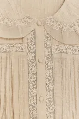 Camisa color crema con cuello bobo, manga corta abullonada con elástico y detalle de bordados combinados a tono y pliegues. Cierre frontal con botones forrados.