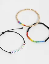 Set de tres pulseras elásticas: una con cuentas blancas con corazones de colores, otra con cuentas negras y detalle de arcoíris, y otra dorada con detalle de arcoíris.