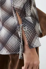 Camisa de manga larga con estampado escocés en tonos chocolate, blanco y gris. Presenta cuello camisero, cierre frontal con lazos y puños con lazos ajustables.