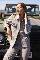 Chaqueta de denim liviana en color beige con detalle de volados en el frente, cuello camisero y bolsillos funcionales. Presenta un calce relajado y cierre frontal mediante botones.