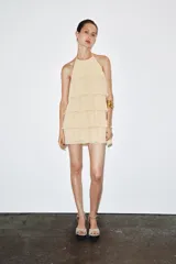 Minivestido color crema con cuello halter ajustable con lazadas y espalda descubierta. Presenta volantes en el mismo tejido.