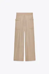 Pantalón de sarga color beige, de tiro medio con cintura elástica en la espalda, bolsillos laterales de plastrón con botón metálico y pierna ancha.