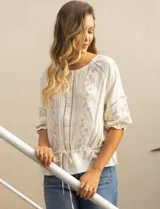 Blusa color crema con cuello redondo, abertura central con botones y mangas 3/4 con puño fruncido. Presenta detalles calados y bordados en mangas, delantera y espalda. La cintura se ajusta con un lazo.