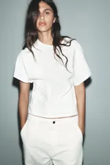 Camiseta blanca de algodón con cuello redondo y manga corta. Presenta bordados florales a tono en el lateral derecho del pecho y en la parte inferior izquierda.