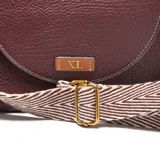 Bandolera pequeña de material sintético color bordó, con textura granulada. Presenta una solapa frontal con costuras decorativas blancas y un logo metálico con las letras "XL". Incluye una correa ancha de tela con diseño a rayas blancas y marrones, ajustable y desmontable, y herrajes metálicos dorados.