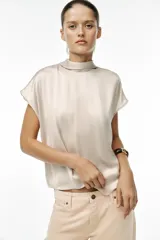 Blusa color crudo de tejido satinado, con cuello subido, manga sisa y cierre en la espalda con lazada y botones.