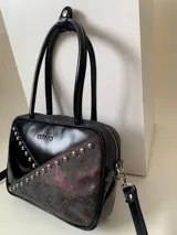 Cartera rígida negra de cuero con tachas plateadas y tres combinaciones de cuero diferentes que aportan textura. Tiene herrajes y cierre metálico plateado marca YKK. Cuenta con un asa fina para llevar al hombro o en la mano y una correa larga extra regulable.