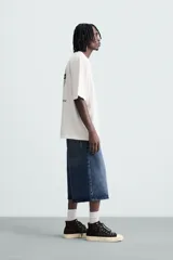 Camiseta blanca de corte holgado, cuello redondo y manga corta. Estampado frontal y trasero con gráficos y textos combinados en color negro.