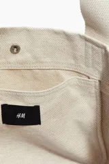 Bolso shopper de lona color beige con doble asa de hombro y cierre de botón a presión. Tiene un compartimento exterior y un compartimento interior pequeño, ambos abiertos.