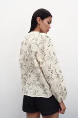 Blusa color crema con bordado floral en azul, de cuello redondo con cordón ajustable y mangas largas abullonadas con puños elásticos.