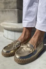 Mocasín náutico dorado de cuero con detalle de lazo en la parte delantera y suela de goma color caramelo.
