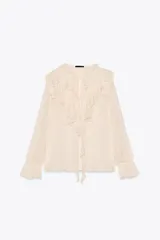 Blusa color crema semitransparente con cuello redondo con volantes y cordón. Manga larga acabada en elástico y volante. Cierre frontal con botones.