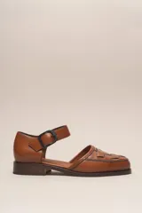 Zapato marrón de cuero vacuno trenzado a mano, con correa al tobillo con hebilla negra y suela de cuero.