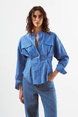 Camisa de manga larga color azul con finas rayas verticales blancas. Presenta cuello mao, botones frontales y dos bolsillos de parche con tapa en el pecho. La prenda tiene un diseño ajustado en la cintura con pinzas y un volumen holgado en las mangas.