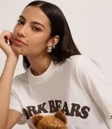 Remera blanca de algodón con cuello redondo y manga corta. Presenta un estampado central de un oso de peluche marrón vistiendo un buzo negro con el texto "NEW YORK" en blanco.