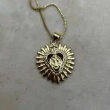 Collar con dije de corazón dorado rodeado de rayos y engarzado con strass. El corazón tiene una abertura central que enmarca una figura religiosa (posiblemente la Virgen María o un santo) y está coronado por una pequeña cruz.