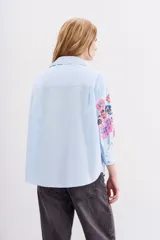 Camisa blanca de algodón con bordados florales y lentejuelas en las mangas, cuello camisero, mangas largas con puños abotonados y corte amplio.